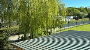 A metal roofed structure, possibly a pavilion, with a clear view of a weeping willow tree and a building in the background.