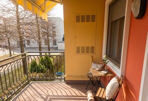 Ein Balkon mit gelber Wand, einem Fenster mit Jalousien, einem kleinen Tisch mit Stühlen und einem Blumentopf.