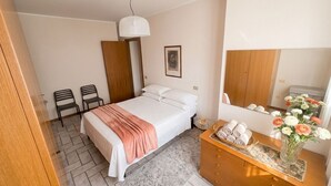 Quarto com cama, duas cadeiras, cômoda com toalhas, espelho e luminária suspensa.