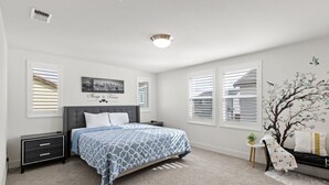 Un dormitorio con cama, mesitas de noche, una lámpara de noche, un cuadro en la pared y una ventana con persianas.