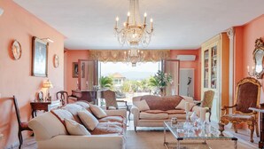 Uma sala de estar ampla, com um lustre grande, vários sofás, uma mesa de centro e vista para o exterior.