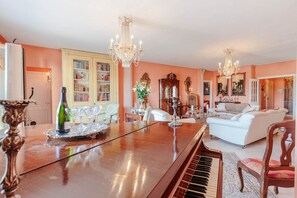 Un vaste salon comprenant un piano, un canapé blanc, une table à manger surmontée d’un lustre et une bibliothèque.