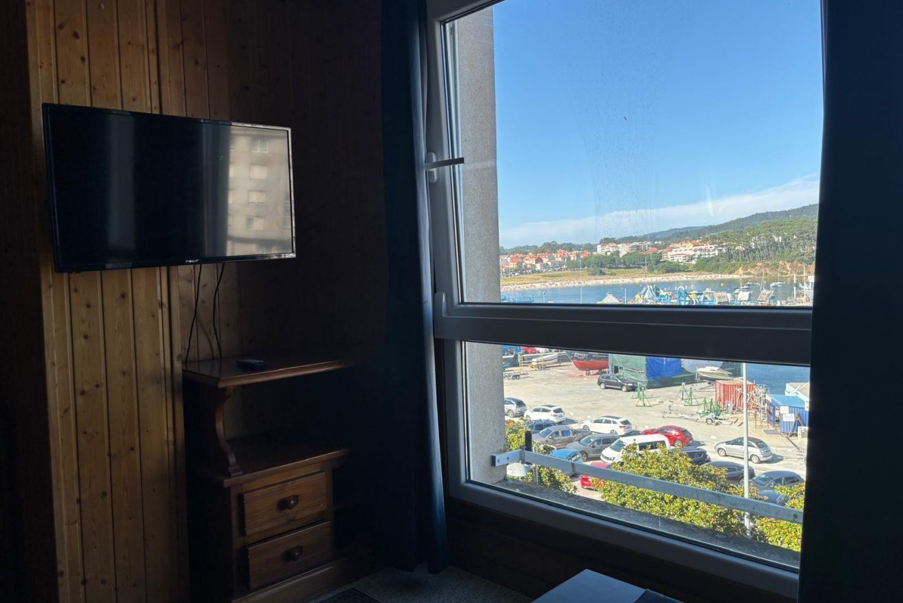 A room with a large window, wooden paneling, a flat-screen TV mounted on the wall, and a small wooden dresser.