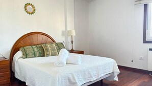 Un lit bien fait, avec un drap blanc et une serviette enroulée, une tête de lit en bois, une lampe de chevet et un miroir fixé au mur.