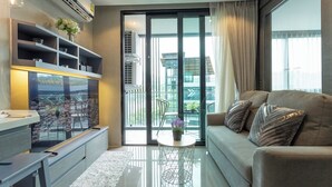 A modern living room with a sofa, a coffee table, a TV, and a balcony with a view of buildings. - Rooftop Pool Condo Walk to Beach, Gym, Wi-Fi c118 (Tambo kamala)