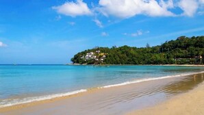 A beach with clear turquoise water, a sandy shore, and a hill with buildings in the background. - Rooftop Pool Condo Walk to Beach, Gym, Wi-Fi c118 (Tambo kamala)