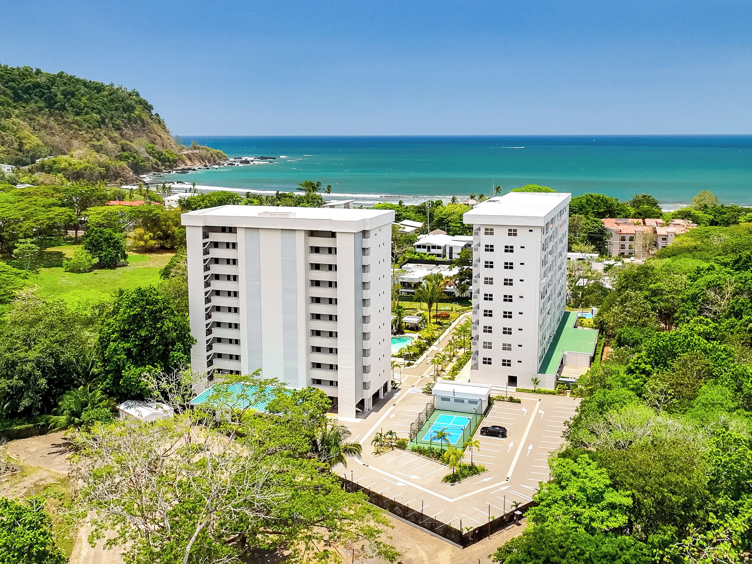 Un hotel de gran altura con piscina y cancha de tenis, rodeado de vegetación y vistas al océano.