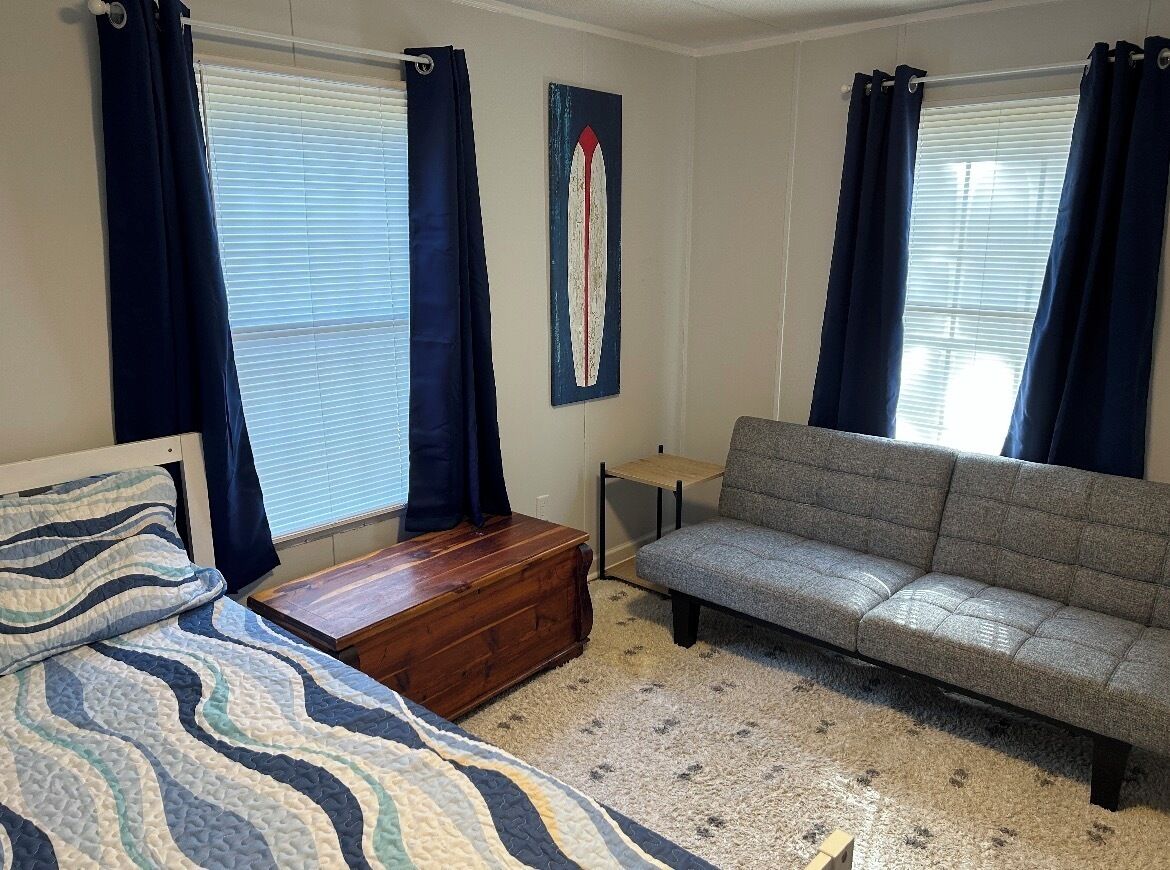 A bedroom with a bed, a grey sofa, a wooden chest, and a small table.