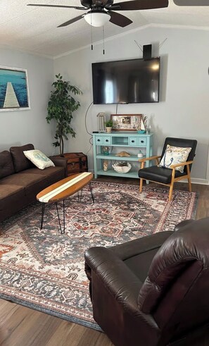 A living room with a sofa, a bench, a chair, a TV, a ceiling fan, and a decorative rug. - Terrific 2-bedroom mobile home,  perfect for bringing your boat (Wilmington)