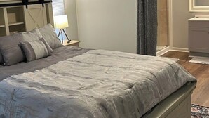 A bedroom with a bed, a ceiling fan, a nightstand, a lamp, a window with blinds, and a bathroom visible through an open door. - Terrific 2-bedroom mobile home,  perfect for bringing your boat (Wilmington)