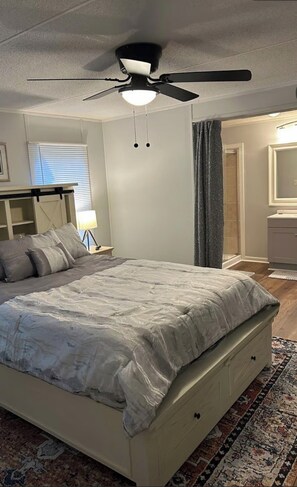 A bedroom with a bed, a ceiling fan, a nightstand, a lamp, a window with blinds, and a bathroom visible through an open door. - Terrific 2-bedroom mobile home,  perfect for bringing your boat (Wilmington)