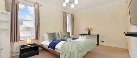 A bedroom with a bed, a bedside table, a lamp, a radiator, a window with curtains, and a television on the wall.