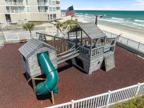 A beachside playground with a slide, wooden play structures, and a view of the ocean. - Shark Tooth Cove: Oceanfront + Pools | Sleeps 8 (North Topsail Beach)