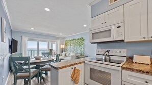 A modern kitchen with white cabinetry, a built-in oven, microwave, and a toaster. There is a dining area with a table and chairs, and a view of the outdoors through large windows. - Skytide | Oceanfront Balcony + Pool & Beach Access (North Topsail Beach)