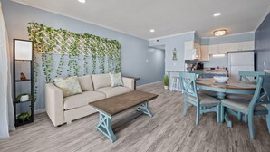 A living room with a sofa, coffee table, and dining area with chairs and a table. - Skytide | Oceanfront Balcony + Pool & Beach Access (North Topsail Beach)