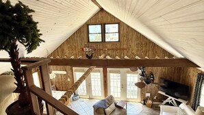 Zona d'estar espaiosa d'estil loft amb un sostre alt, parets de fusta, una finestra gran i una escala que mena a un nivell superior.