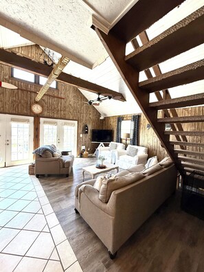 A spacious living room with a high ceiling, wooden beams, a large TV, and a sofa. - Memory Maker: Cozy 3-bedroom house in quaint Oak Island steps to the beach (Oak Island)