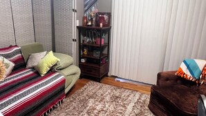 Ein Wohnzimmer mit einer Couch, einem braunen Sessel, einem Bücherregal und einem Fenster mit Jalousien.
