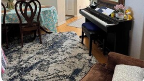 Ein Wohnzimmer mit Klavier, einem Esstisch mit Stühlen, einem Teppich und einer Küche im Hintergrund.