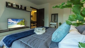 A bedroom with a bed, a television, a shelf with decorative items, and a door leading to another room. - Beach Walk Condo Poolside Retreat by the Sea C123 (Tambon Rawai)