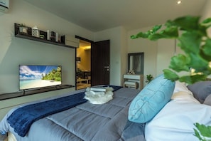 A bedroom with a bed, a television, a shelf with decorative items, and a door leading to another room. - Beach Walk Condo Poolside Retreat by the Sea C123 (Tambon Rawai)