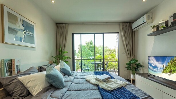 A modern bedroom with a large bed, a TV, a balcony with plants, and a view of greenery. - Beach Walk Condo Poolside Retreat by the Sea C123 (Tambon Rawai)