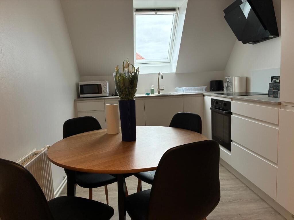 A compact kitchen with a microwave, sink, and stove. There's a round wooden table with chairs, a potted plant, and a skylight above.