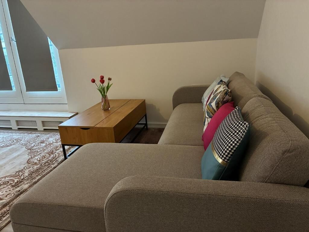A living room with a sofa, a small wooden table, a vase with flowers, and a patterned cushion.