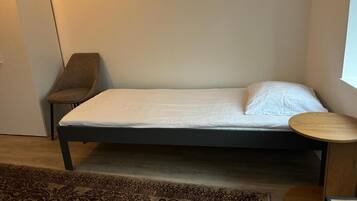 Una cama individual con colchón blanco y estructura oscura, una mesita redonda de madera, una alfombra estampada y una silla.