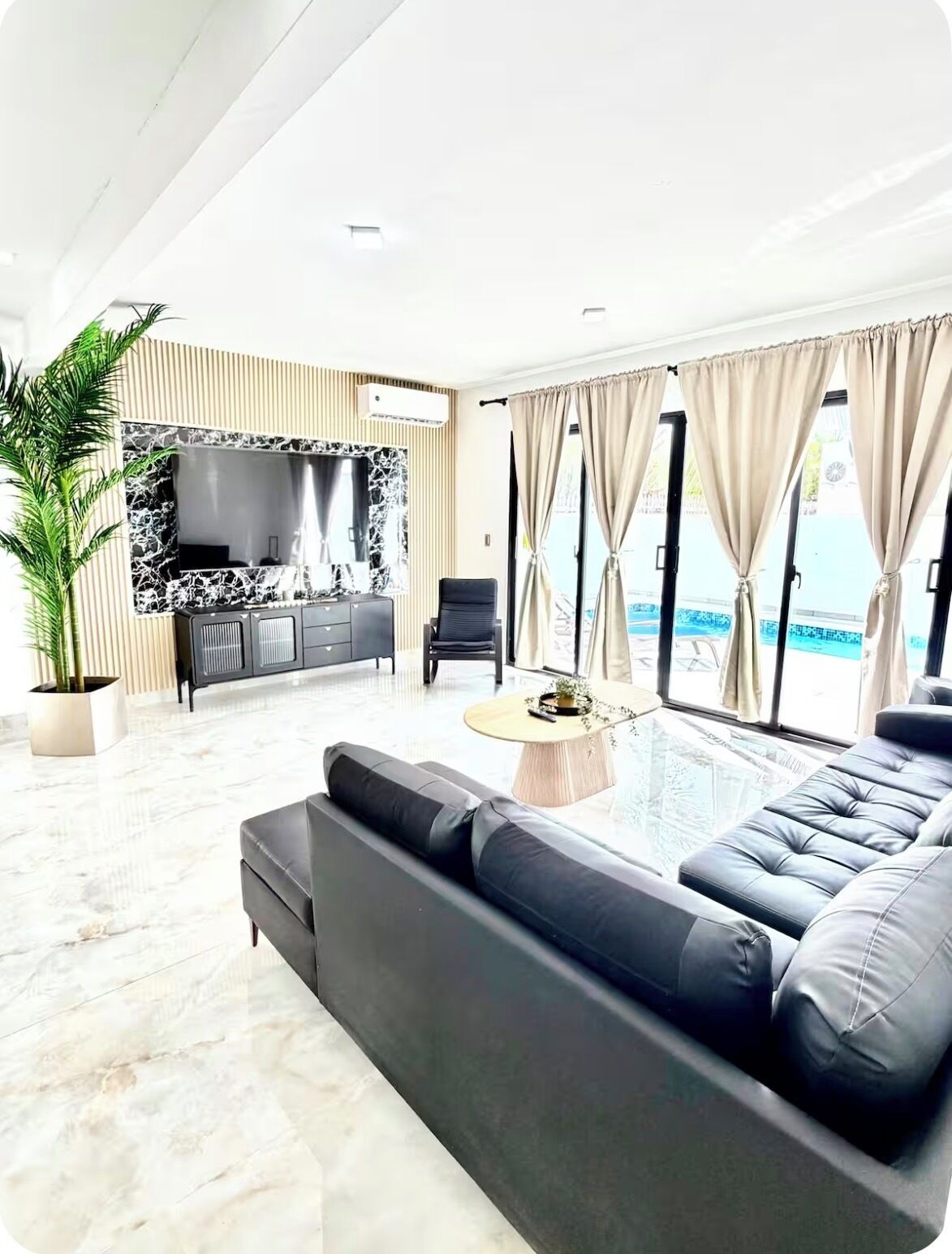 A modern living room with a large black sofa, a coffee table, and a TV against a marble wall. There is a potted plant and a view of a swimming pool through large windows.