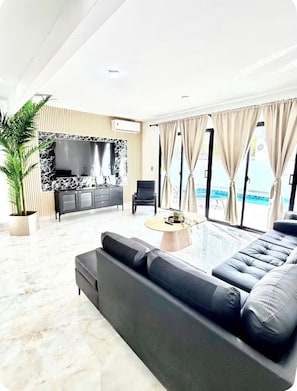 Un salon moderne avec un grand canapé noir, une table basse et un téléviseur placés devant un mur en marbre. On y trouve une plante en pot et, grâce à de grandes fenêtres, on peut admirer une piscine.