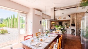 A dining area with a long table set for a meal, a kitchen with a brick oven, and a view of a garden with tall trees. - Luxury Villa With Pool in Lucca (Capannori)