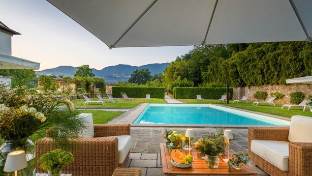 A poolside area with wicker furniture, a table with flowers, and a view of a swimming pool and mountains. - Luxury Villa With Pool in Lucca (Capannori)