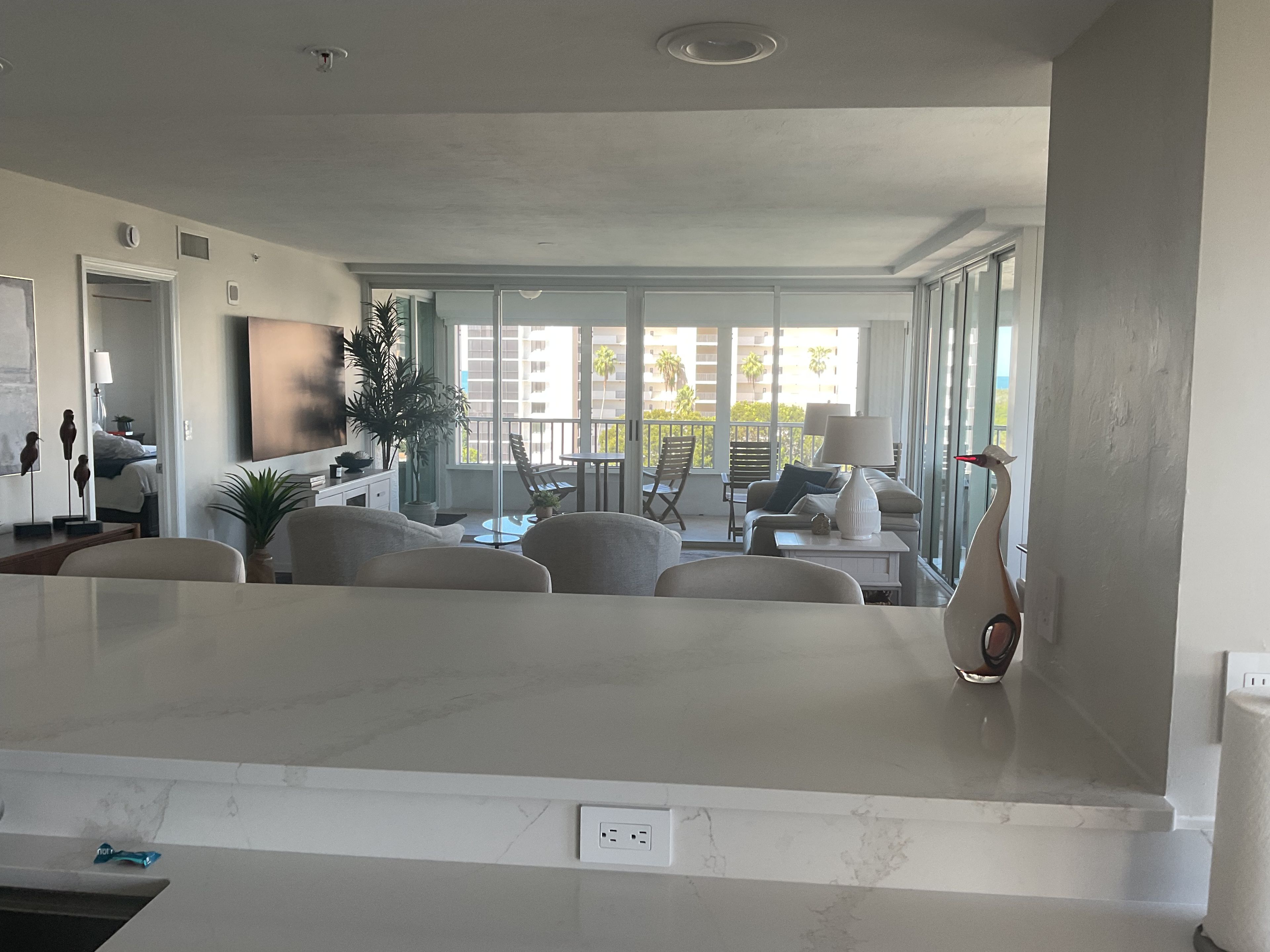 A modern living room with a large white marble countertop, a sofa, a dining table, and a balcony with a view of palm trees.