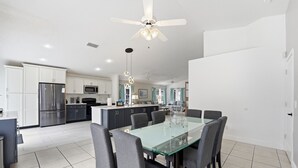 Una cocina moderna con armarios blancos, un refrigerador de acero inoxidable, un microondas y un comedor con una mesa y sillas de vidrio.
