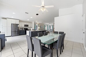A modern kitchen with white cabinets, a stainless steel refrigerator, a microwave, and a dining area with a glass table and chairs. - Spacious 4BR Pool Home Peaceful Views (Kissimmee)