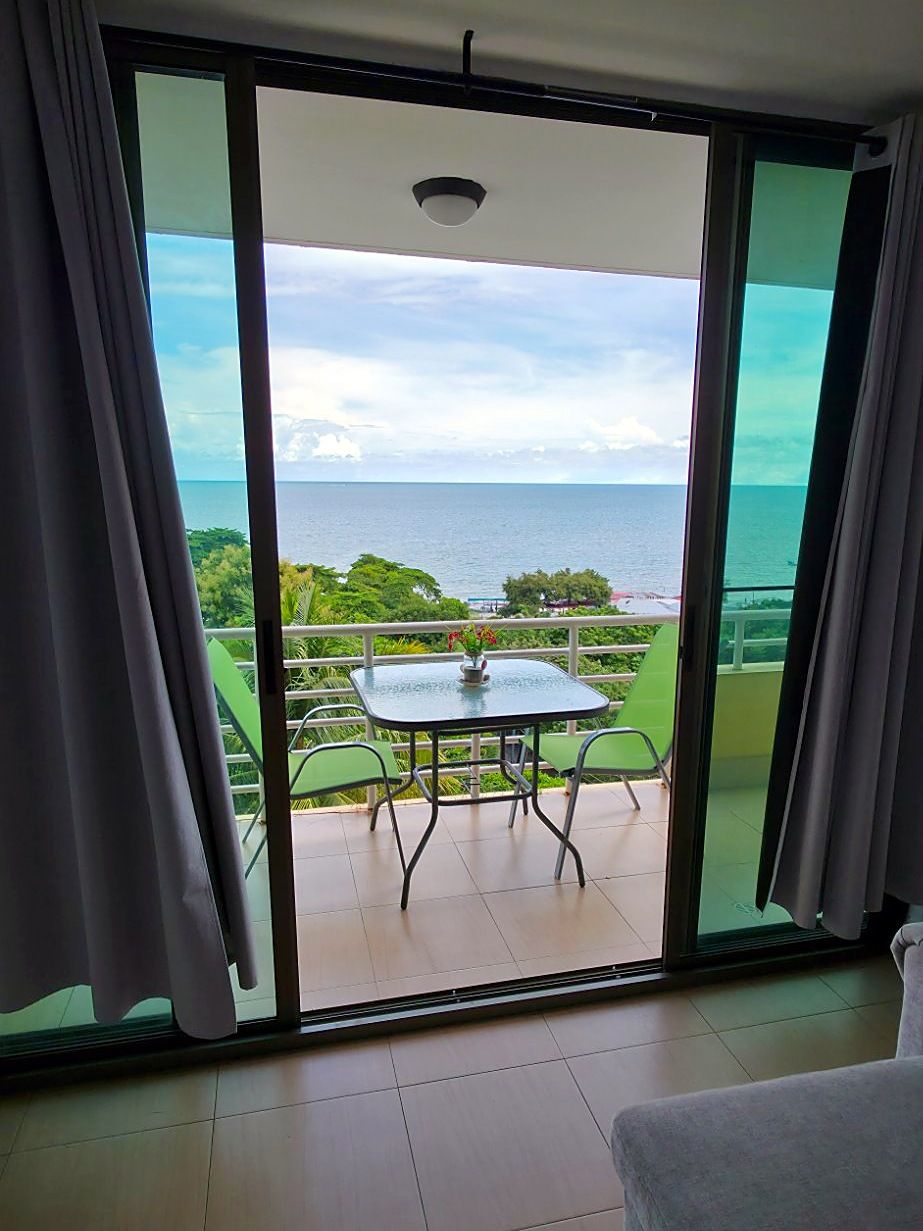 A room with a sliding glass door leading to a balcony with a table and chairs, overlooking a body of water and greenery.