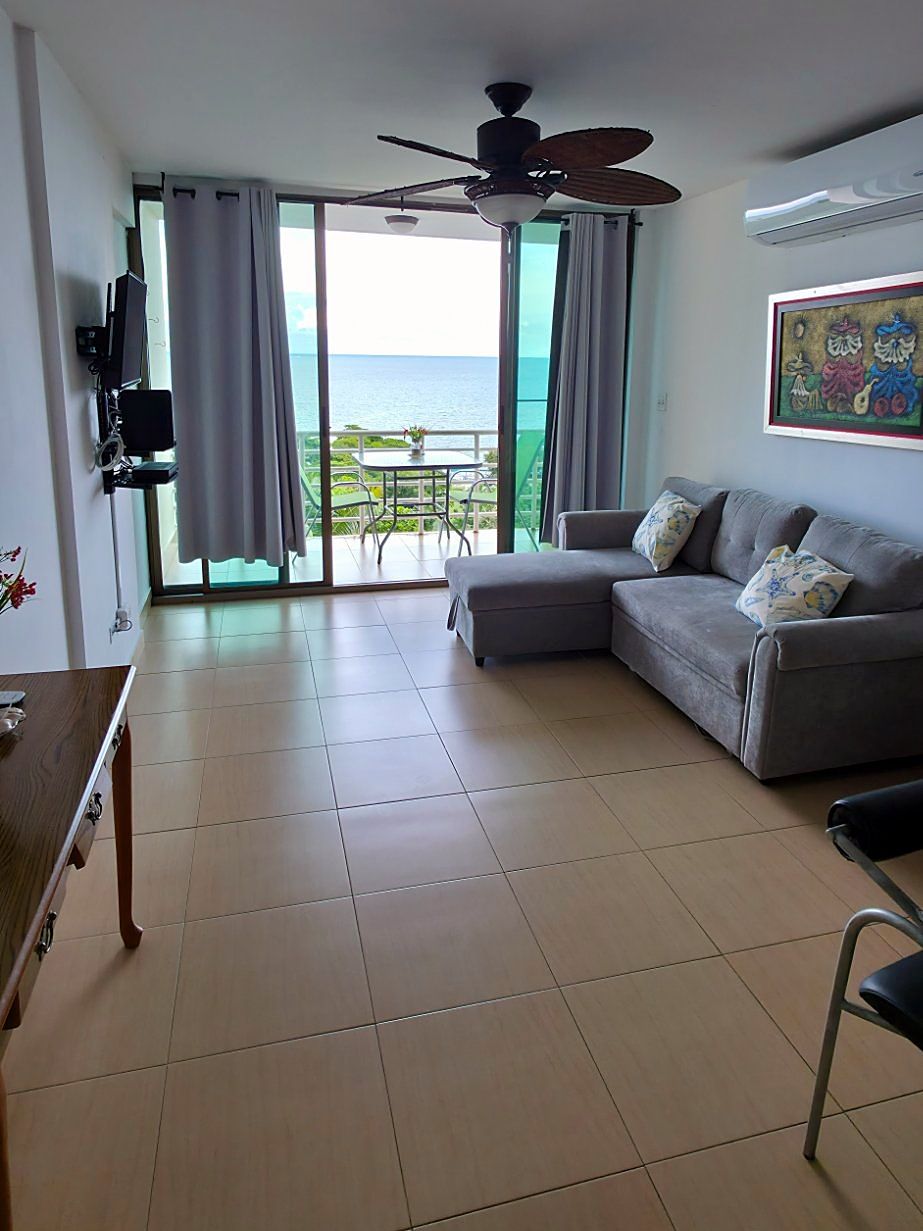 A living room with a sofa, a ceiling fan, a TV, and a view of the sea through large windows.