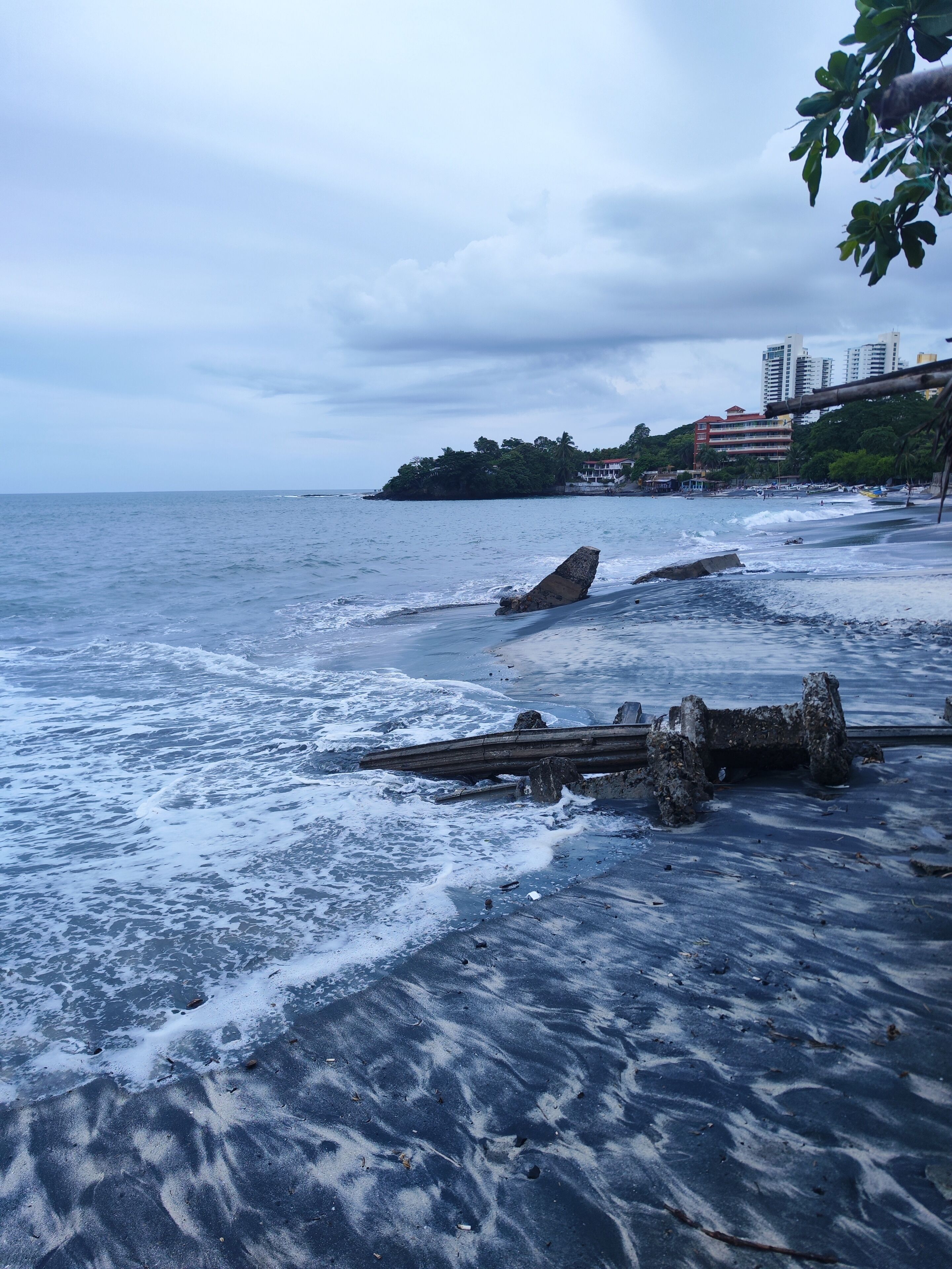 Ein Strand mit Wellen, einer Holzkonstruktion und Gebäuden im Hintergrund.