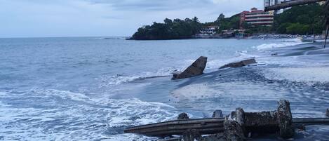 Una playa con olas, una estructura de madera y edificios al fondo.