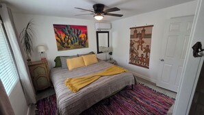 Une chambre à coucher avec un lit, un ventilateur de plafond, un tableau, un miroir, une porte et une fenêtre avec des rideaux.