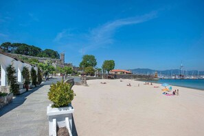 Una playa con una estructura parecida a un castillo, cielo despejado y un puerto deportivo al fondo.