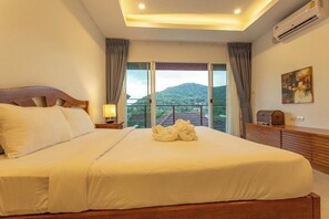 A hotel room with a large bed, a nightstand, a lamp, a wooden dresser, and a view of a mountain through large windows. - 2BR Walk Award Winning Beach Pool & Gym C126 (Tambon Rawai)