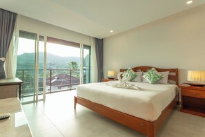 A hotel room with a large bed, a wooden headboard, a nightstand with a lamp, and a balcony with a view of greenery. - 2BR Walk Award Winning Beach Pool & Gym C126 (Tambon Rawai)