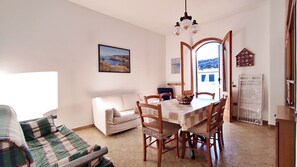 Ein Wohnzimmer mit einer Couch, einem Esstisch mit Stühlen, einem Bild an der Wand und Blick durch eine geöffnete Tür.