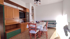Ein Zimmer mit Esstisch, Stühlen, einem Fernseher auf einem Regal und einer Couch.