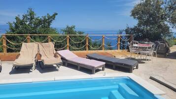 Un área de piscina con sillones de descanso y una mesa, rodeada por una terraza de madera y vistas al mar.