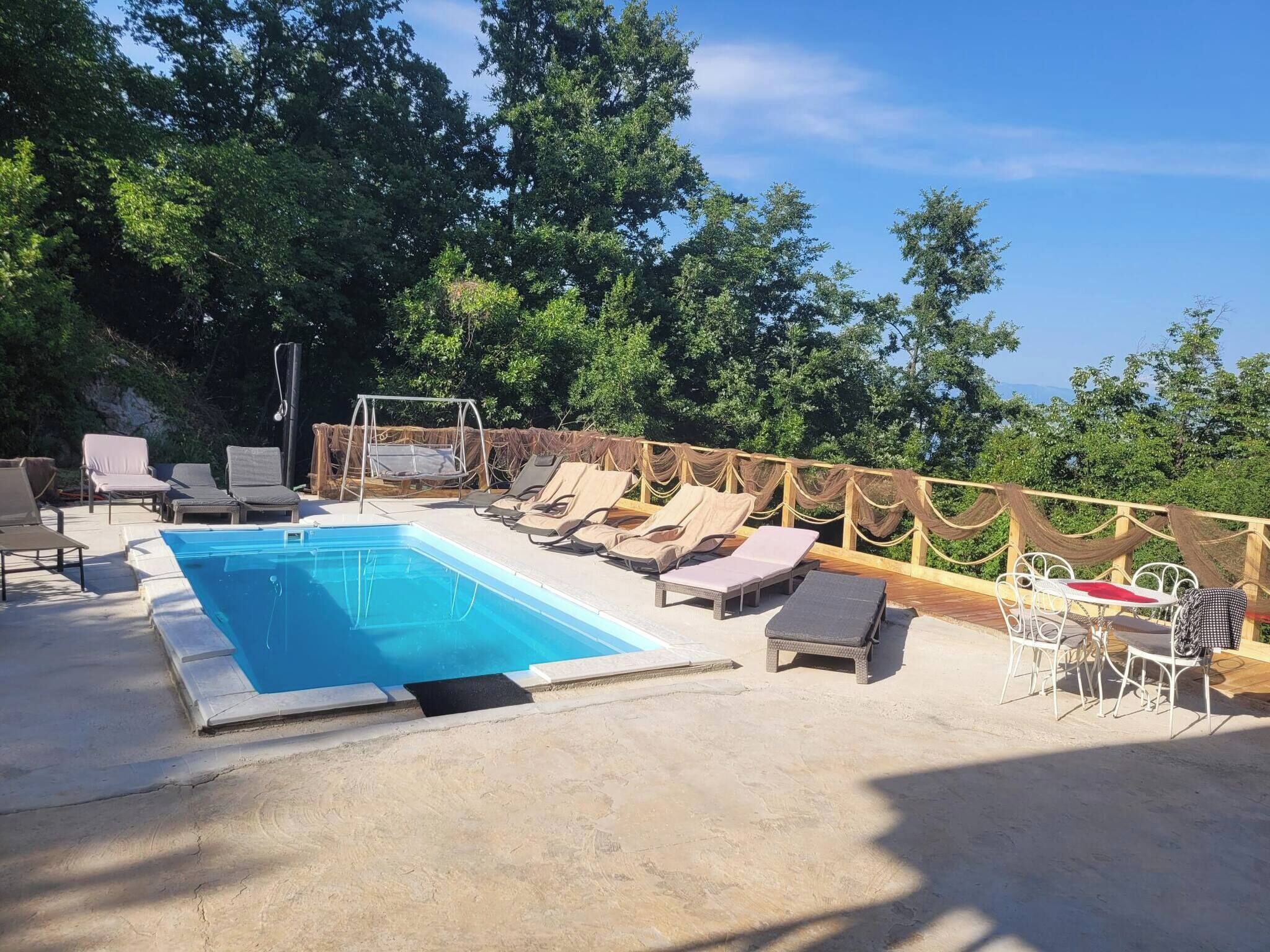 Ein Schwimmbecken mit Liegestühlen und einem Tisch, umgeben von einer Holzterrasse und Bäumen.