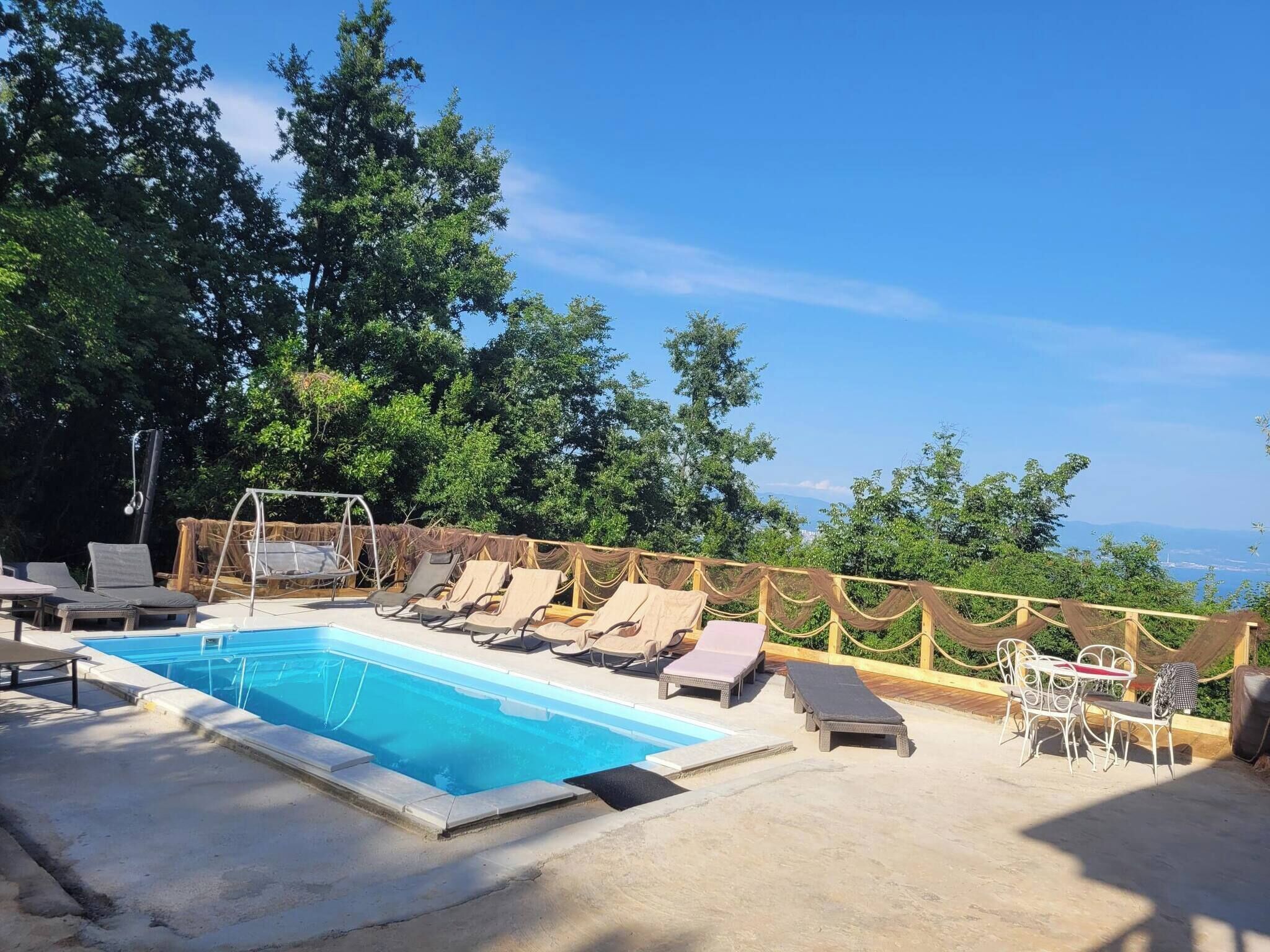 A pool area with lounge chairs and a table, surrounded by trees and a clear sky.