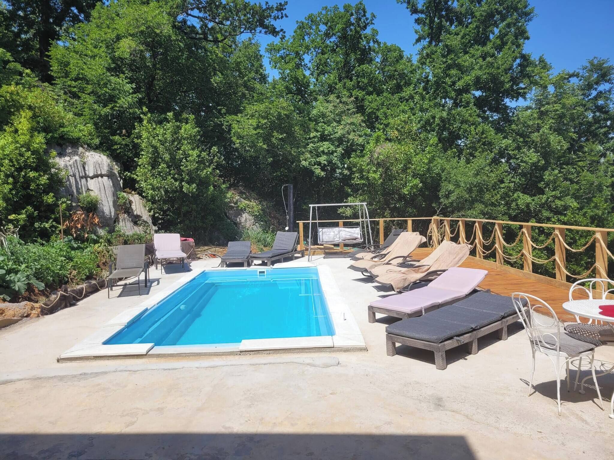 Une piscine rectangulaire entourée d’une terrasse en bois et d’une végétation luxuriante, avec des chaises longues et une table à proximité.